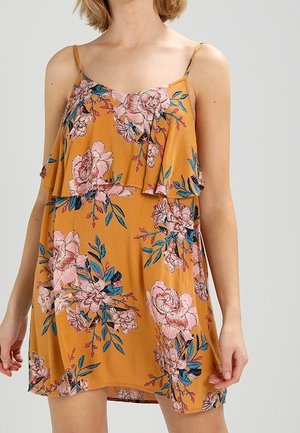 Robe sans manches moutarde avec imprimé floral rose et superposition à volants portée par une personne visible des épaules jusqu'au milieu des cuisses.