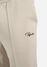 Beige treningbukser laget av teksturert stoff, med et brodert "Pegador"-logo i svart på siden. Inkluderer lommer og elastisk midjebånd.