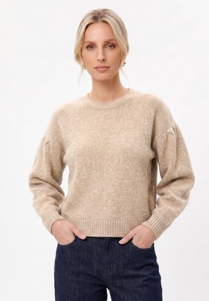 Femme blonde portant un pull beige en tricot et un jean bleu foncé, debout les mains dans les poches sur un fond blanc uni.