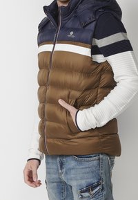 Padded vest with a hood, featuring a brown body and navy upper, white stripe, front zipper, and side pockets. White long-sleeve shirt visible underneath.