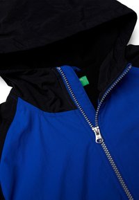 Niebiesko-czarna kurtka zapinana na zamek z kapturem, o gładkiej fakturze, kontrastowym przeszyciu oraz zieloną metką wewnątrz kołnierza.