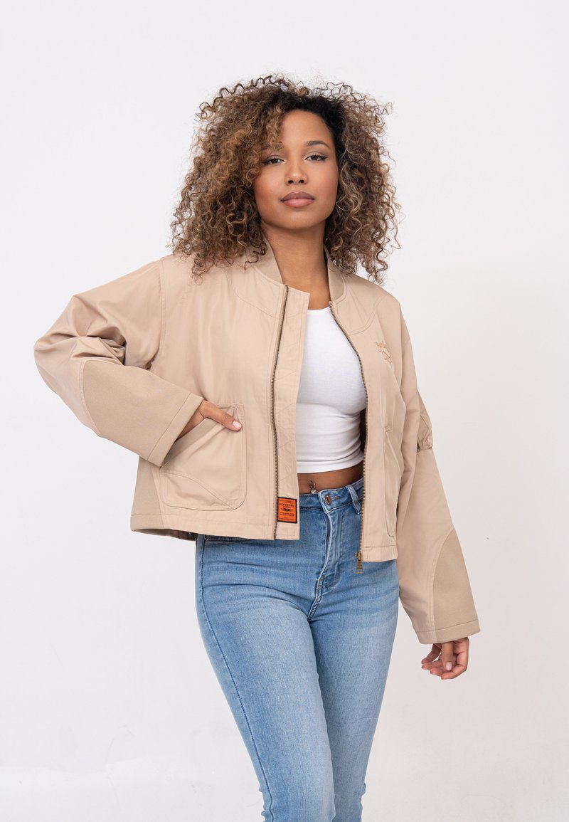 Veste cropped beige avec fermeture éclair à l'avant, manches larges et logo brodé. Dispose de poches latérales et d'un tissu texturé avec des accents lisses.