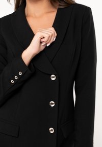 Donna con blazer nero e bottoni argentati che aggiusta il risvolto, mostrando parte del collo e della mano con unghie curate.