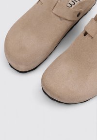 Beige Wildleder Clogs mit runder Zehenpartie, schwarzen Sohlen und einem seitlichen Riemen mit Knopf. Weiche Textur mit minimalistischem Design und ohne sichtbare Muster.