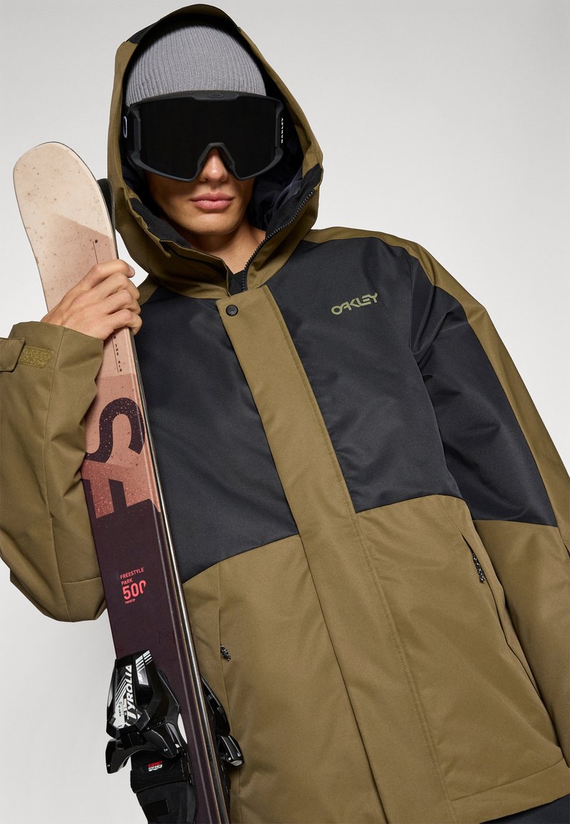 Veste de ski en vert olive et noir, avec une texture lisse et une capuche réglable, assortie à un masque de ski noir et à un ski avec fixations.