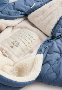 Gesteppte blaue Außenschichtjacke, gefüttert mit weichem cremfarbenem Fleece. Verfügt über ein Etikett mit Pflegehinweisen und cartoonhaften Akzenten. Mit Reißverschluss.