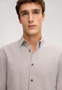 Camicia button-up grigia chiara con tessuto testurizzato, colletto morbido e bottoni marroni. Presenta una vestibilità comoda e sottili righe.
