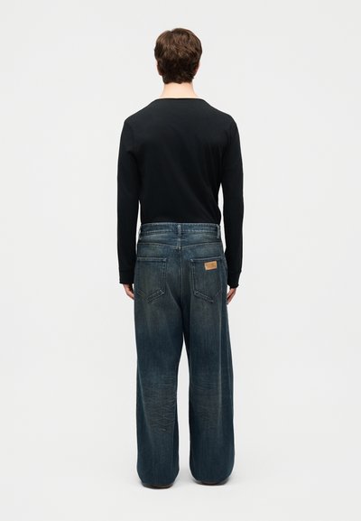 Un homme se tient de dos, portant une chemise noire à manches longues et un jean bleu délavé, ample, avec des poches arrière et une étiquette de marque en cuir.