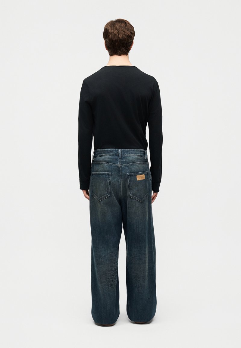 Man staat met de rug naar de camera, gekleed in een zwart shirt met lange mouwen en loszittende, vervaagde blauwe jeans met achterzakken en een leren merkembleem.