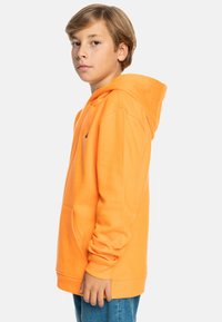 Sudadera naranja con capucha, con un bolsillo frontal y puños de canalé. Textura suave, corte relajado, combinada con jeans azules.