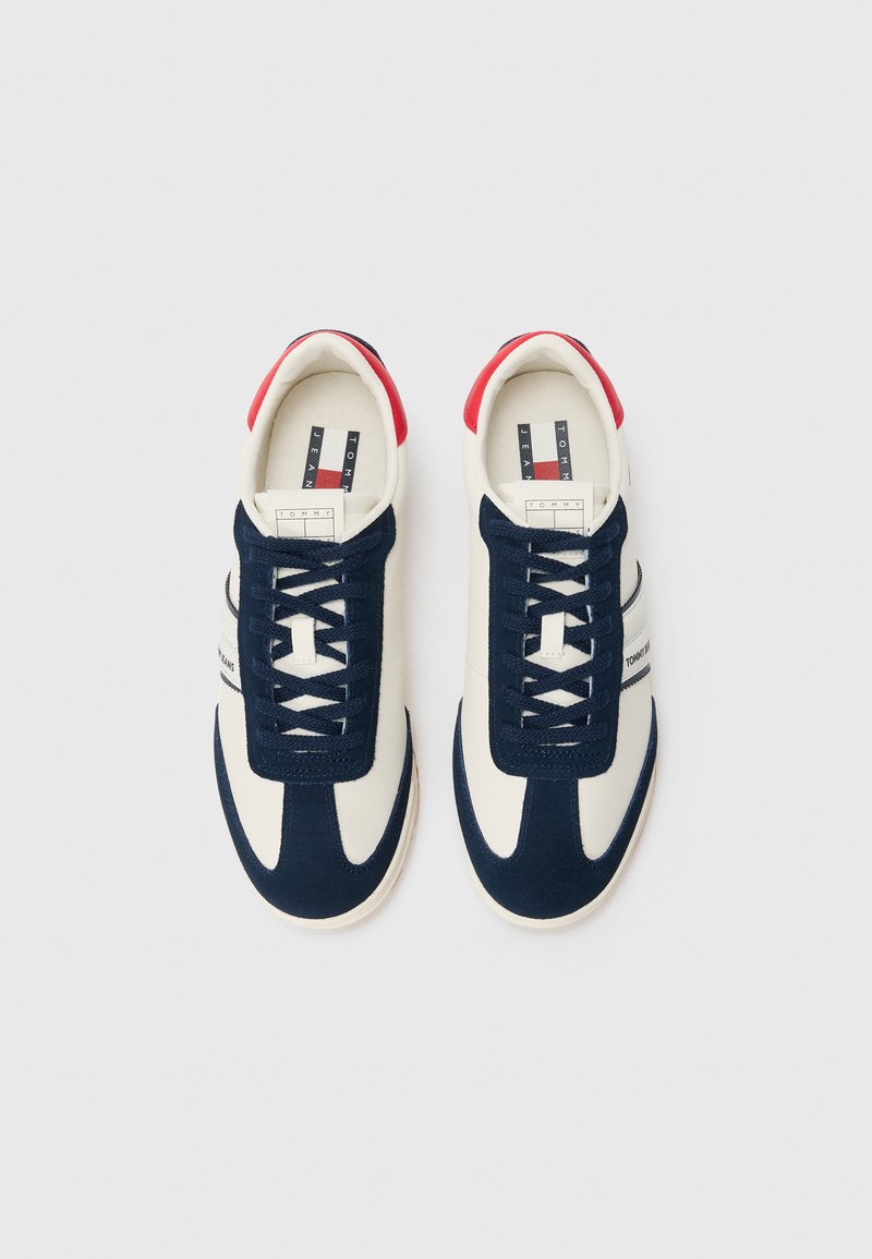Une paire de baskets blanches et bleu marine avec des accents rouges au niveau du talon et des lacets noirs, déposées côte à côte sur un fond blanc.
