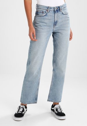 Femme portant un jean droit taille haute bleu clair avec des ourlets bruts et des baskets noires et blanches, debout sur un fond blanc.