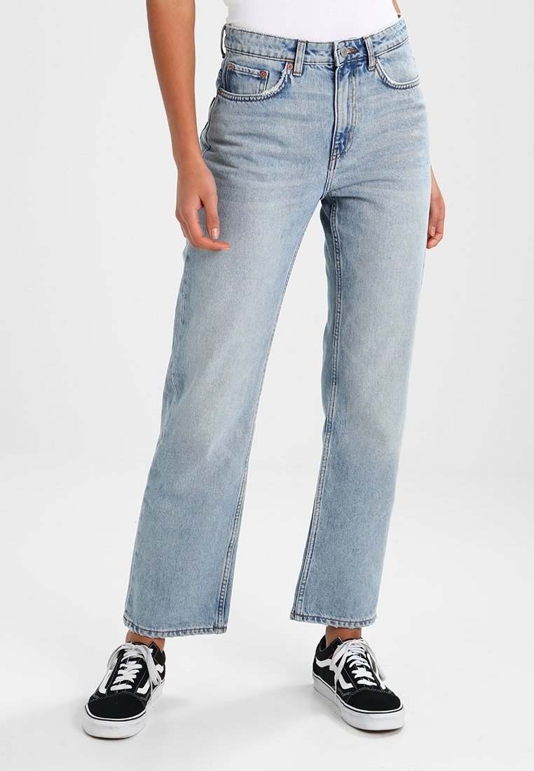 Femme portant un jean droit taille haute bleu clair avec des ourlets bruts et des baskets noires et blanches, debout sur un fond blanc.