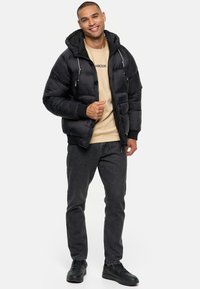 Sort pufferjakke med hætte, der har lynlåslommer og ribbet kant, båret over en lys beige skjorte og mørke jeans. Sorte sneakers.