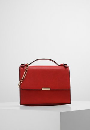 Sac à main en cuir rouge avec anse supérieure et bandoulière chaîne dorée reposant sur un piédestal blanc sur fond uni.