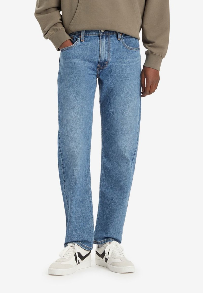 Pantalones vaqueros de mezclilla azul claro con un diseño de pierna recta, cinco bolsillos y ligero desgaste. Combinados con zapatillas blancas y grises.