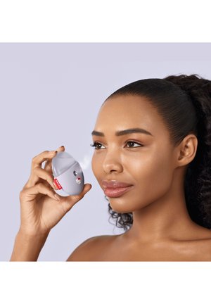 Femme utilisant un petit spray facial à brume porté à la main près de sa joue avec un fond violet doux.