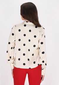 Blusa crema con pois neri, maniche lunghe, polsino con bottoni e colletto alto. Tessuto satinato morbido e vestibilità rilassata.