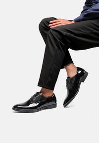 Scarpe in vernice nera con finitura liscia, design con lacci, dettagli in rete e tacco basso, abbinate a pantaloni scuri.