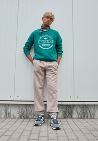 Felpa verde con logo e dettagli bianchi, pantaloni della tuta beige e sneakers grigie con strisce bianche. Sullo sfondo si vede un muro testurizzato e dell'erba.