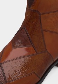 Τμήμα καφέ δερμάτινης μπότας με γεωμετρικά μπαλώματα που ράβονται, με λείες, ερπετικές και υφασμάτινες δερμάτινες υφές πάνω σε μαύρη σόλα.