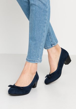 Pieds chaussés de souliers en daim bleu foncé à petits talons avec des nœuds, associés à un jean bleu clair retroussé, debout sur un sol clair.