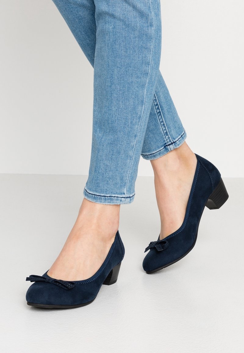 Pieds chaussés de souliers en daim bleu foncé à petits talons avec des nœuds, associés à un jean bleu clair retroussé, debout sur un sol clair.
