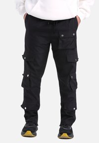 Schwarze Cargo-Hosen mit mehreren Taschen, Druckknopfverschlüssen und verstellbarem Kordelzugbund. Hergestellt aus strapazierfähigem Material, kombiniert mit strukturierten Sneakers.