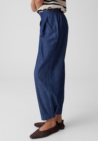 Femme portant un pantalon en denim bleu ample avec une taille élastique et des ballerines marron, debout devant un fond clair uni.