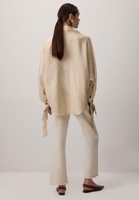 Bluza crem cu model, cu manșete supradimensionate, asortată cu pantaloni evazați de culoare albă murdară și pantofi slip-on maro. Material neted și fluid.