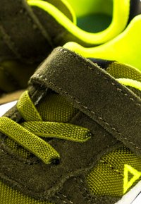 Primo piano di una sneaker verde scuro e gialla neon con lacci e una fascia a strappo su uno sfondo luminoso.