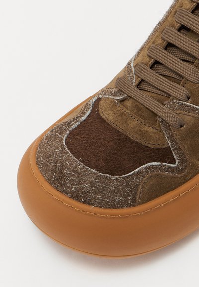 Adidași maro cu panouri din suede texturat și fetru. Prezintă o talpă din cauciuc de culoare bej cu vârf rotund și șireturi maro. Cusăturile sunt vizibile de-a lungul marginilor.