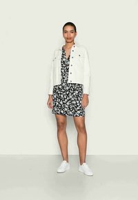 Jaqueta de ganga branca sobre um vestido floral preto. O vestido apresenta um decote em V, e a modelo usa ténis brancos, em pé contra um fundo neutro.