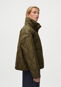 Casaco puffer acolchoado em verde oliva com gola alta, bolsos laterais e uma silhueta rectângulo. Apresenta um padrão texturizado e ondulado no tecido.