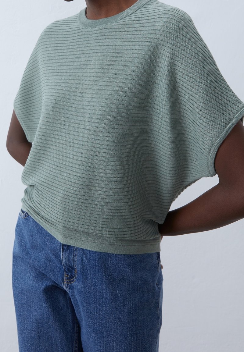Personne portant un haut en tricot à manches courtes côtelé vert clair, associé à un jean en denim bleu taille haute, les mains derrière le dos.
