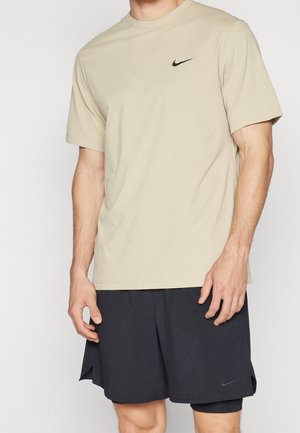 Muž v béžovém tričku Nike a černých vrstvených šortkách Nike stojí před jednoduchým světlým pozadím.