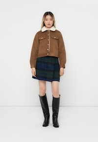Vrouw in een bruine jas met sherpa-kraag, blauwe en groene geruite rok, en zwarte kniehoge laarzen, staande tegen een effen witte achtergrond.