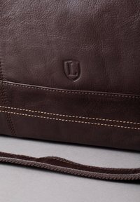 Brown leather bag featuring a smooth texture, stitched accents in lighter thread, and an embossed logo on the lower corner.