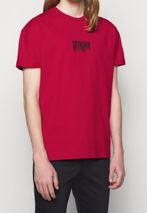 Person trägt ein rotes T-Shirt mit kurzen Ärmeln und aufgekrempelten Bündchen sowie schwarzem Schriftzug "DRYKORN" auf der Brust, kombiniert mit schwarzen Hosen.