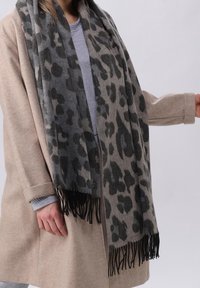 Personne portant un manteau beige et un pull gris avec une grande écharpe à imprimé léopard noir et gris avec des extrémités frangées.