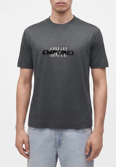 Homme portant un t-shirt à manches courtes gris foncé avec le logo "EMPORIO ARMANI" en noir et blanc, associé à un jean bleu clair.