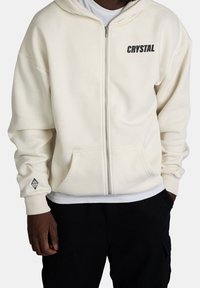 Felpa zip con cappuccio color crema, dalla vestibilità rilassata, con tasche frontali e la scritta "CRYSTAL" stampata in nero sul petto. Include il logo a forma di diamante ricamato sulla manica.