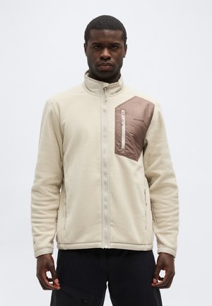 Hombre con chaqueta de forro polar beige con bolsillo en el pecho color malva con cremallera y pantalones negros, de pie contra un fondo blanco liso.