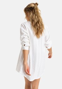 White, textured shirt with long sleeves rolled to the elbows, featuring button details. Falls to mid-thigh, showcasing a relaxed fit.