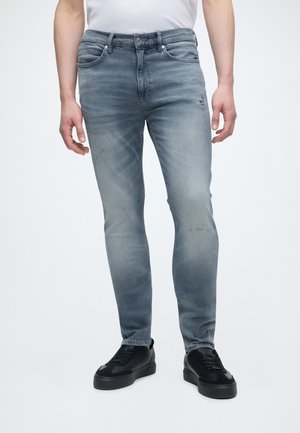 Άνδρας με ξεθωριασμένο γκρι τζιν στενής γραμμής και μαύρα αθλητικά παπούτσια στέκεται σε απλό λευκό φόντο.