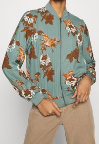 Chaqueta bomber azul claro con un patrón floral y de pájaros en marrón y rosa. El diseño incluye un cierre de cremallera y puños acanalados.
