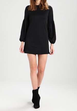 Kobieta w czarnej mini sukience z długimi bufkami i czarnych botkach do kostek stoi na jednolitym jasnym tle.