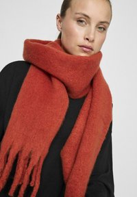 Écharpe en laine rouge avec une texture douce et duveteuse, dotée de bords frangés. Portée lâchement autour du cou contre un haut sombre.