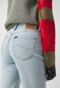Gros plan sur une personne portant un jean bleu clair avec une étiquette marron "Lee", assorti à un pull en tricot multicolore, mettant en valeur les détails de la tenue.