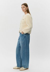 Krémová, štruktúrovaná bunda s okrúhlym výstrihom, zapínaním na gombíky a dvoma náprsními vreckami, kombinovaná so svetlomodrými širokými nohavicami a krémovými teniskami.